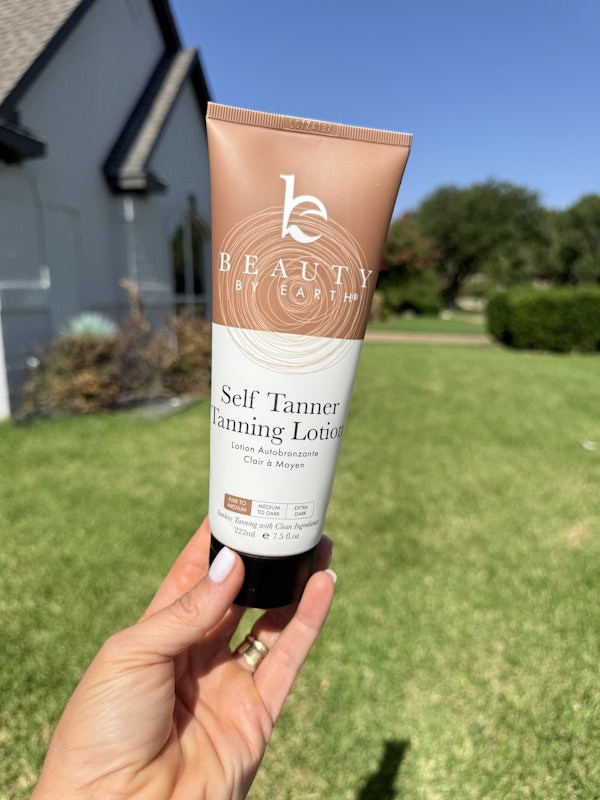 Hand holding a tube of 'Beauty by Earth' self Tanner in an outdoor setting.