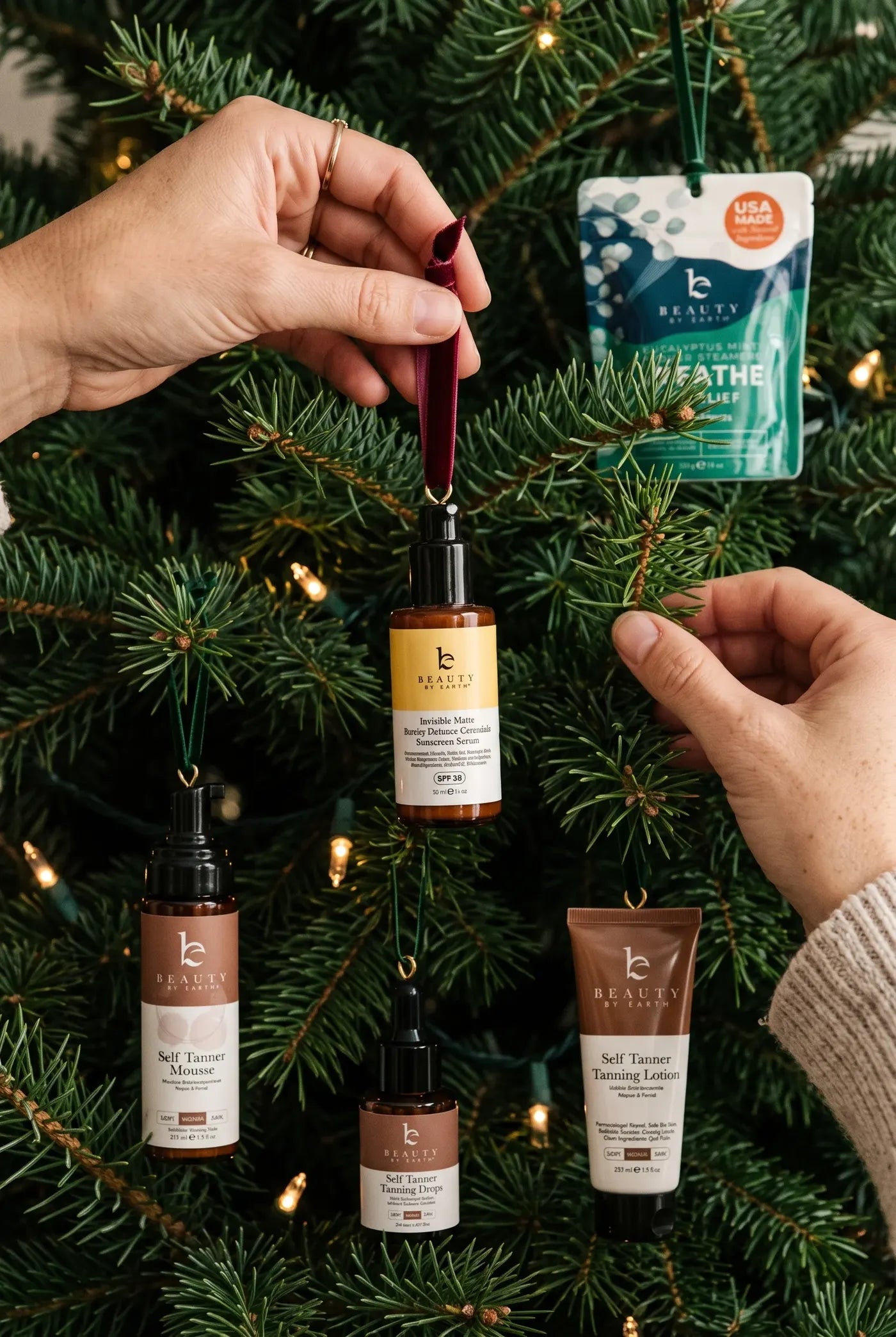 Hands hanging Beauty by Earth products as ornaments on a lit Christmas tree
